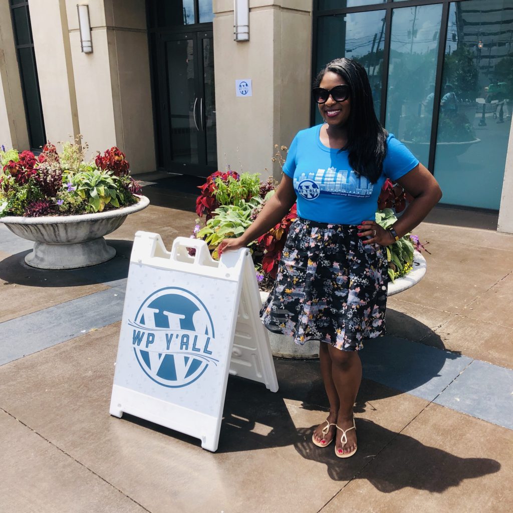 An African-American woman (me!) dressed in a blue t-shirt and floral skirt poses in front of a building standing next to a sign that reads "YP Y'all"